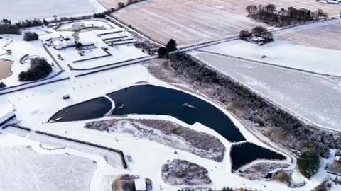 PA Media Overhead shot of a fish-shaped lake surrounded by fields which have a light dusting of snow and ice on them. 