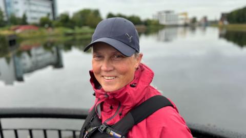 Woman standing on a bridge over a body of water. She is wearing a bright pink waterproof jacket, a black cap and a black life vest. She is smiling.