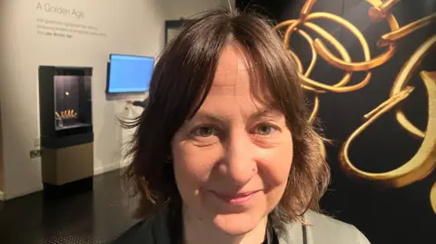 Niamh Baker who has short brown hair and is standing in a museum. She is wearing a grey suit jacket and has a stud piercing in her nose. 