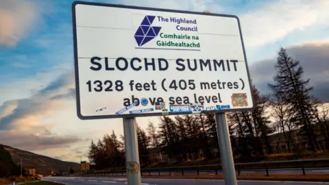 A large road sign on the A9. It has Highland Council's name in English and Gaelic and the words "Slochd Summit" and its height above sea level - 1,328ft (405m).