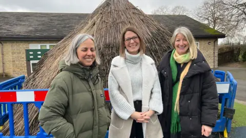 Three women in front of a roundhouse in a playground. They are all smiling at the camera and wearing padded winter coats