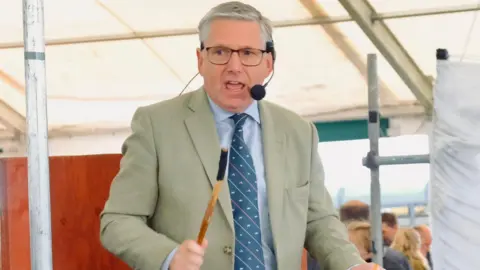 Scott Donaldson Scott Donaldson is a clean-shaven man of about 60 wearing a green-grey jacket and blue shirt and tie. He is presiding over an auction and is speaking through a head mounted microphone. He holds a stick used to signify a sale in his hand.
