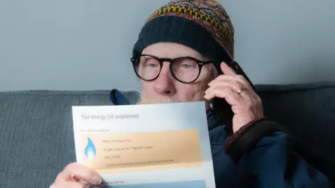 Getty Images Older man sits on sofa wearing woolly hat and speaking on the phone while holding energy bill