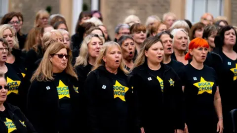 PA Media A group of women wearing black T-shirts with yellow Rock Choir T-shirts. They are singing outside a stone building.  One is wearing sunglasses while another has orange hair.