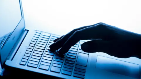 A laptop computer viewed from the side. Part of the screen is visible, while a hand is typing on the keyboard