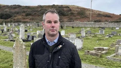 Adrian Hughes standing facing the camera in a daytime picture, wearing a dark coat and a blue checked shirt.  He's in Llandudno's Great Orme cemetery surrounded by green grass punctuated by a diverse range of mainly white gravestones, with a brown bracken-covered hill behind.