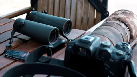 A pair of binoculars and a camera with a long lens are on a table. A chair is next to the table. 