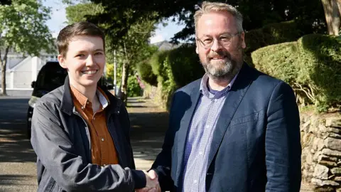 Dylan Tippetts in an orange shirt and blue jacket shaking hands with chairperson of Plymouth Liberal Democrats, Stuart Bonar