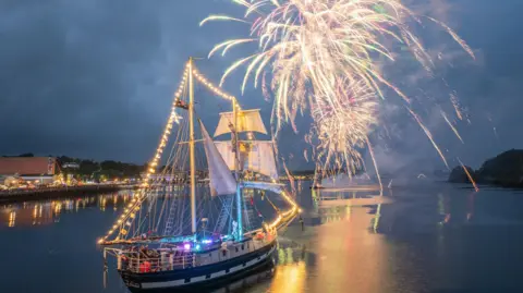 DCSDC fireworks go off above the river Foyle , a tall ship is in the foreground. It is dark and the ship is lit up 