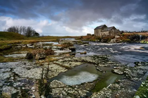 John Cuthbert The stone mill building at Hawick can be seen across a river, with frozen puddles of water between the rocks in the foreground. 