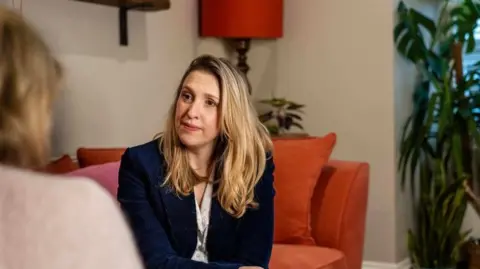 Clementine Wade A woman with long blonde hair sat on an orange sofa, she is wearing a dark blue blazer and white blouse and is in conversation with a woman whose face is not visible, she has brown hair and is wearing a beige jumper. 