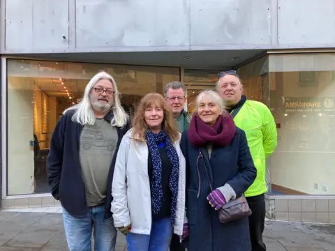 Three men and two women group together wearing winter clothes in front of an empty shop