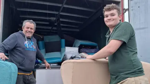 Chris James Baird and his son, standing by a lorry, with items in it and smiling for the camera. James to the left is wearing a blue jumper and his son is in a green polo shirt.
