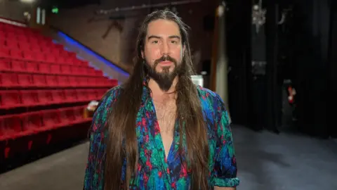 Adam looks at the camera as he wears a bright shirt which is unbuttoned down his chest. Adam has long dark brown hair which comes past his chest and a beard. Behind him is rows of theatre seats in the Arts Centre