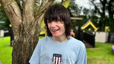 A young man stands in a garden. He has dark shoulder length hair with a heavy fringe. He is smiling widely and wearing a blue t-shirt. behind him is a bright sunny garden, with grass and treats and some play equipment. 