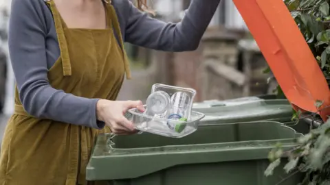 A woman wearing a khaki jumpsuit opens a green bin with an orange lid. She is holding a plastic tray filled with some other plastic rubbish over the open bin. The background is out of focus, but she is on a residential street.