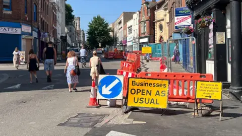 A street in sunshine with orange barriers and metal signs on the right hand side near the shops, with people walking along the road up towards a large tree in the distance.