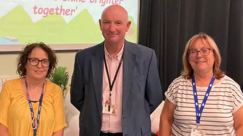 Joan Maguire had mid-length brown hair and is wearing dark framed glasses and a yellow top. Jeremy Barnes is wearing a blue suit jacket and a pink and white striped shirt. Clair Fealey has mid length brown hair and is wearing dark framed glasses and a white t-shirt with horizontal black stripes. All are stood alongside each other in a school hall smiling at the camera.