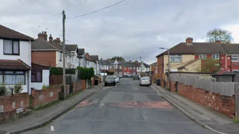 A residential street of red brick houses on both sides of a road, with some shrubbery visible and garden walls and fences. There is a red "20" speed limit sign painted onto the surface of the tarmac. 