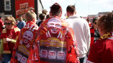 EPA Views of fans walking to the stadium with flags draped on their back, showing Arne Slot and a slot machine. The flags say Arne's slot machine