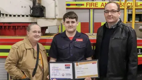 Robert in fire cadet uniform holding a certificate in front of a fire engine. Either side of him are his parents.