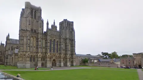 The front of Wells Cathedral with the green in foreground and people walking along paths