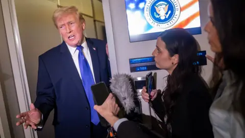 Getty Images Donald Trump gestures as he speaks to reporters while wearing a suit and blue tie and holding onto a door frame next to a screen that has the president's logo on it that says 'seal of the president of the United States', on Air Force One on Monday.