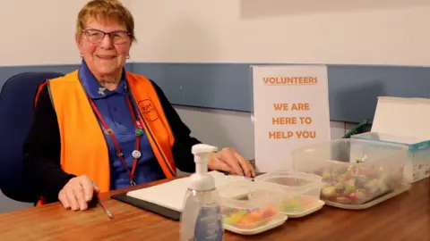 BBC Margaret Wildgust, wearing a hi-vis vest, sat behind a desk which has boxes of sweets on it and next to a sign saying 'we are here to help you'