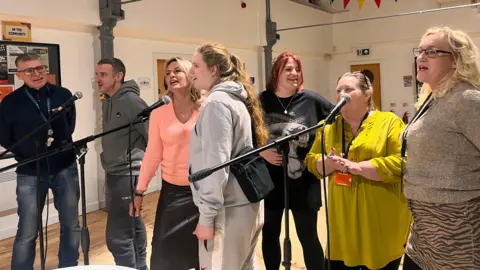 Seven people sing in front of microphones.