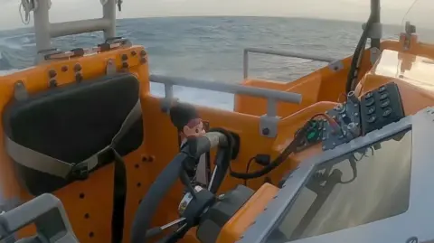 Supplied An elf wearing a black costume appears to be driving an orange lifeboat at sea.