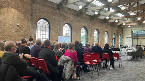 Shariqua Ahmed Rows of people seated on red chairs in a large room. They are facing committee members, who are seated in an oval-shaped arrangement. 