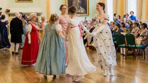 Jane Austen Festival A group of four women wearing regency era dresses is dancing in a circle while holding hands.