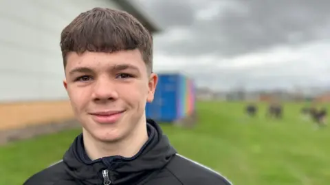 Harley is pictured in a tracksuit on a playing field at school. He has dark brown hair and is in his mid-teens. 