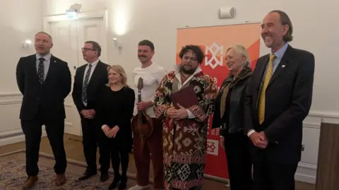 Seven people - made up of five government officials, an artist and a musician - stand in a room with white walls in front of an orange sign. The government officials are all wearing smart clothing. One of the artists has a white T-shirt on and is holding a musical instrument. The other artist is wearing a cloak with various patterns on it.