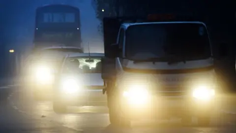 EPA A truck, with two cars behind it have bright headlights in the early morning light