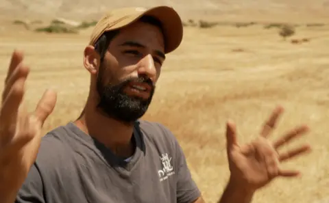 A man with a black beard is being interviewed in a dart coloured t-shirt and beige cap