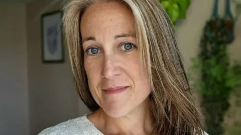 Poppy Whiteside smiling slightly at the camera with her home, with a framed piece of art and plants on the wall, in soft focus in the background