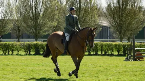 Countryside Lincs A man riding a bay-coloured horse in a field. The man is wearing a black helmet, white jodhpurs, long black boots, a green tweed jacket, a white shirt and a dark-coloured tie. The horse's mane is plaited and it is wearing a bridle and a saddle. It is galloping.