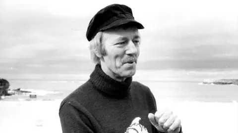 Rick Stevens/Fairfax Media via Getty Images A black and white image of Horace Dobbs. He is standing on a beach and is wearing a black cap and knitted jumper. 