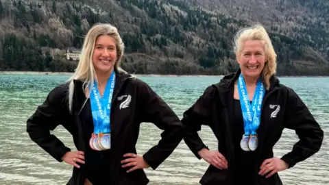 Frozen.ferns/Instagram Two blonde women in black wet suits with medals around the necks stand with their hands on their hips in front of a lake smiling at the camera.