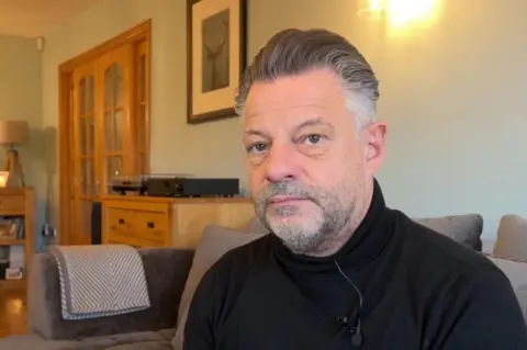 Chief executive of Darlington FC, David Johnston, who has short greying hair and a trimmed beard and is wearing a black high-necked jumper. He is pictured on his sofa at home with a picture on the wall and a pine chest of drawers behind him.