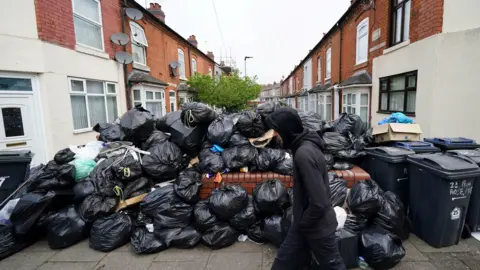 PA Media A man in a black hoodie walks past black bin bags piled up on a residential street, with terraced houses on either side.