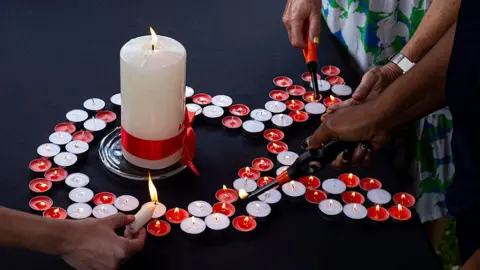 Small red and white candles are arranged in the shape of an Aids ribbon. A large white candle is in the centre. The candles are being lit by some disembodied hands. 