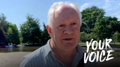 BBC A man with receded grey hair in a blue and white striped shirt stands in front of a boating pond with just a little water left in it and trees in the background
