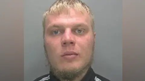 A police custody photo of Eimantas Zemeliauskas. He has short blonde hair and a wispy beard. He is wearing a black top with a white stripe over each shoulder