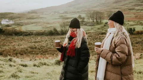 Getty Images Two women walk through the countryside. They both have long blonde hair and wear black beanie hats. One wears a red scarf and one wears a white scarf, and both wear long puffer coats - one in black and the other in camel. Both women carry coffee cups. Behind them is a misty field and a hill.