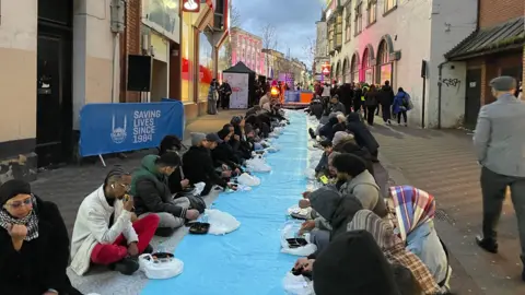 Two lines of people sat across from each other on a city street with a blue carpet between them and each person eating from a prepared meal bag.