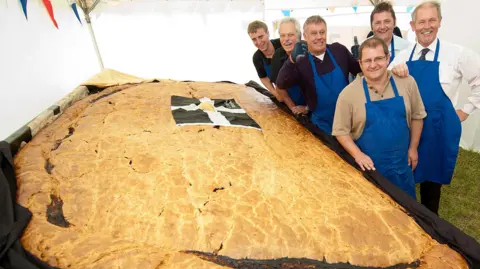 Proper Cornish Pictured is the largest Cornish pasty ever made. There are six workers stood next to the pasty. They are all pictured wearing blue aprons.
