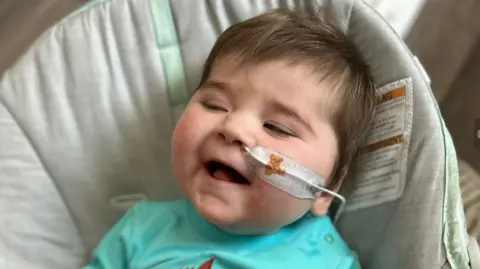 Jack has short brown hair and is wearing a turquoise top and sitting in a baby seat. He has a tube up his nose which is taped to his face with a plaster with a teddy bear on it. He is laughing.