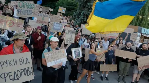 EPA/Shutterstock Dozens of people, mostly young, holding cardboard signs in a protest, as one woman waves a large Ukrainian flag